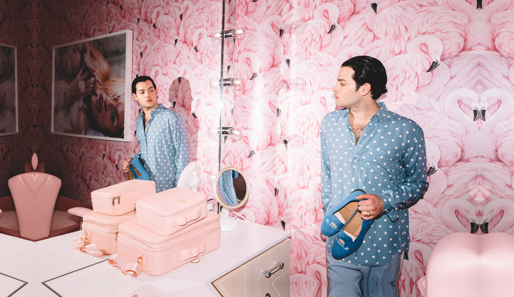 A man photographed in a pink room looking in the mirror holding a pair of blue slippers.